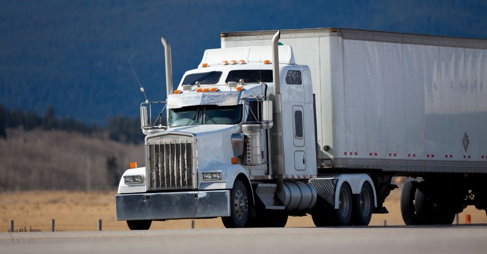 A white semi-truck carrying a trailer on a highway.