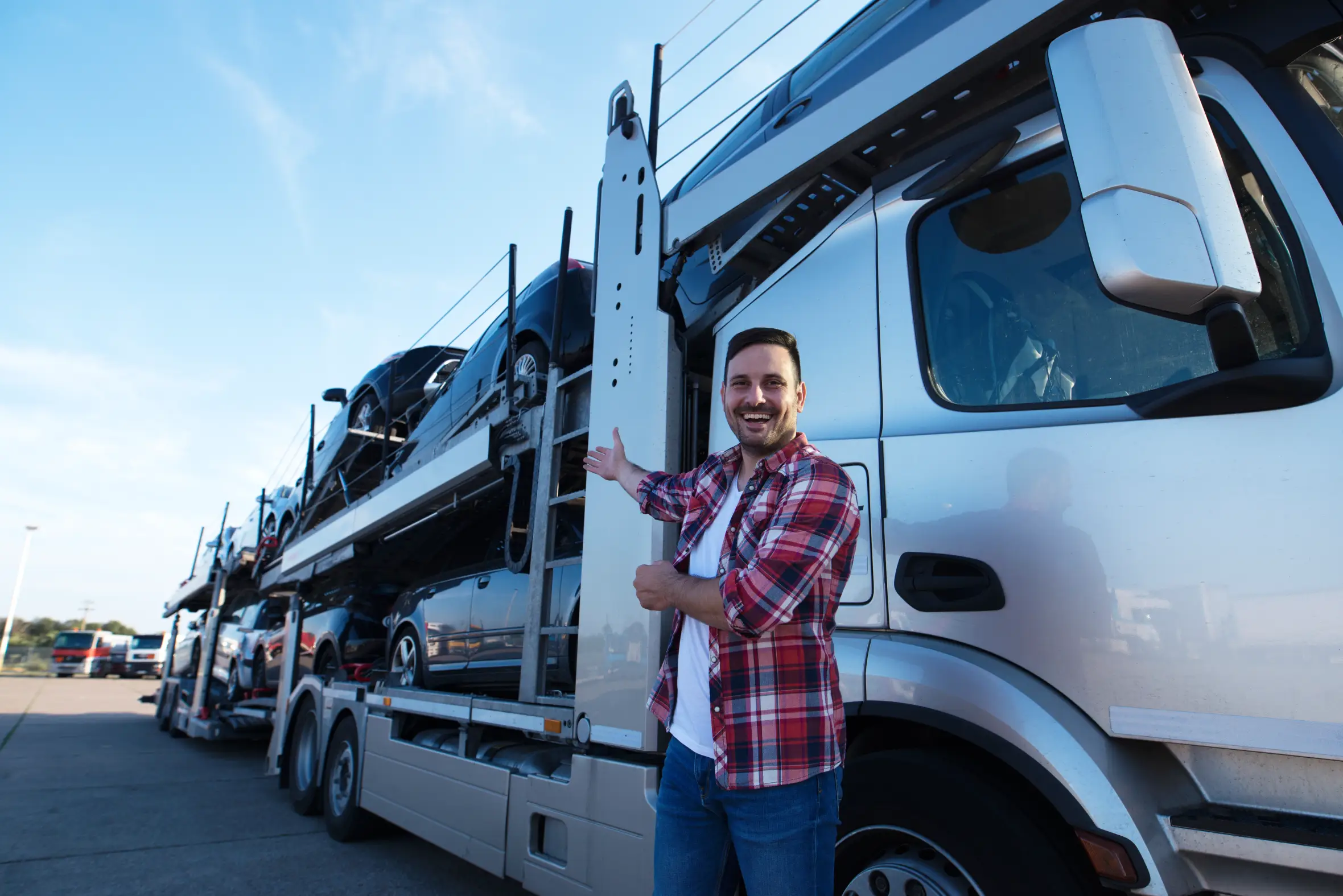 A smiling male truck driver in a plaid shirt and jeans stands next to a car hauler semi-truck loaded with black cars, gesturing towards the truck with his right hand.