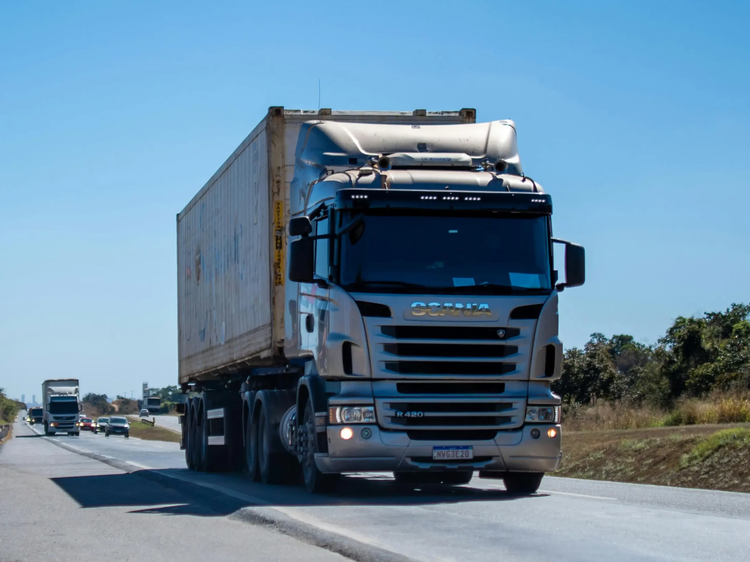 A silver Scania semi-truck with a beige cargo container is driving on a multi-lane highway under a clear blue sky.