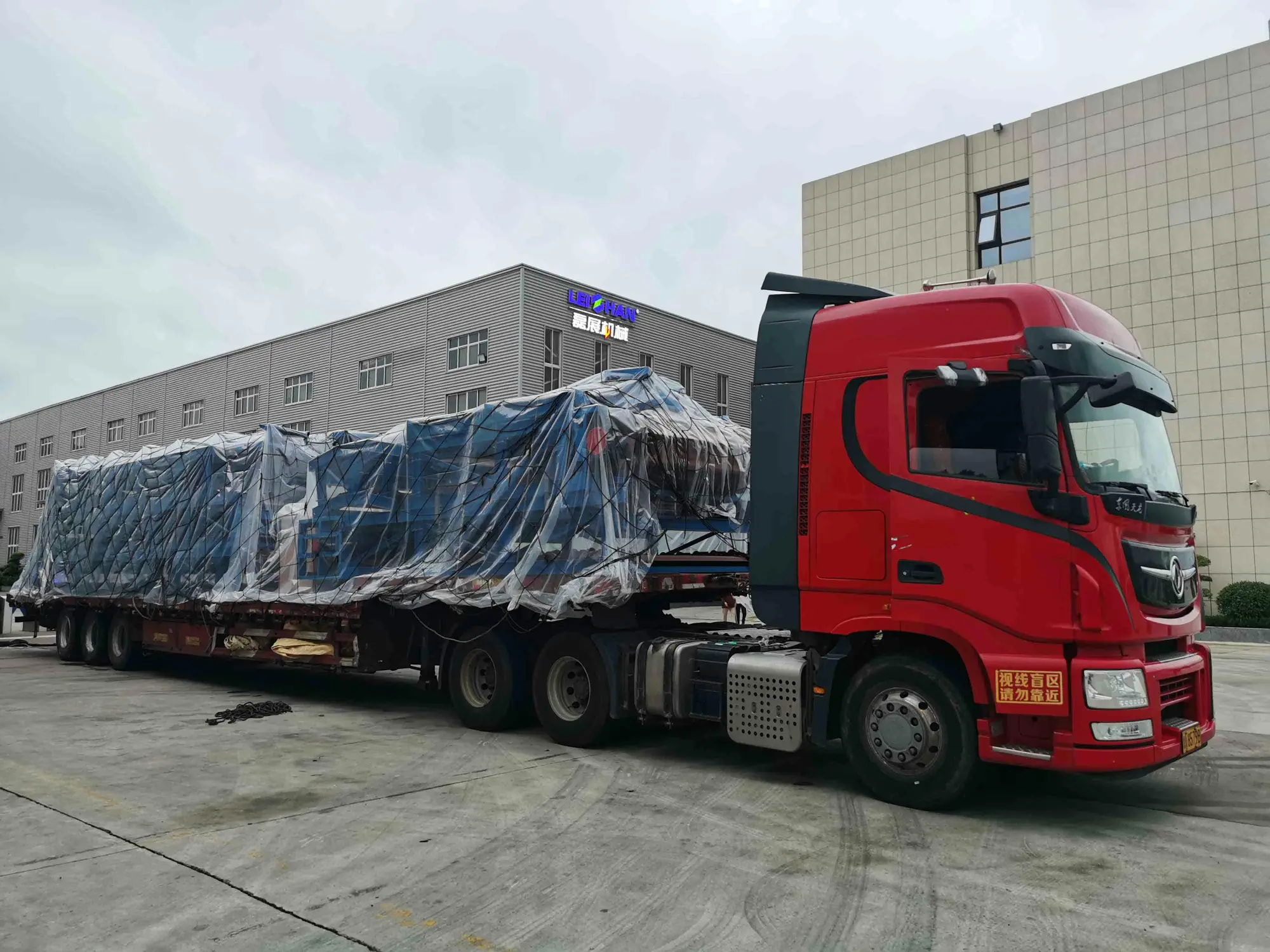 A red semi-truck is parked in front of a building, hauling a large, blue, tarp-covered load on a flatbed trailer.