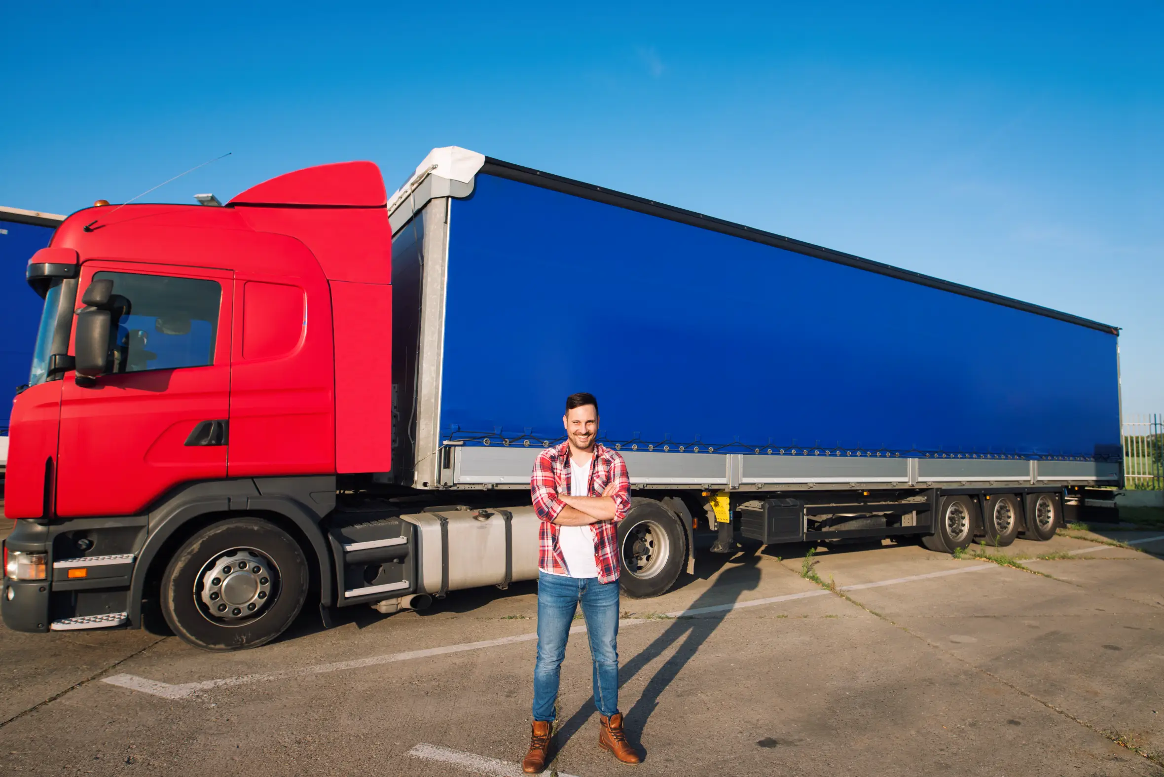 A male truck driver with a beard and plaid shirt stands in front of a red semi-truck with a blue trailer, arms crossed and smiling.