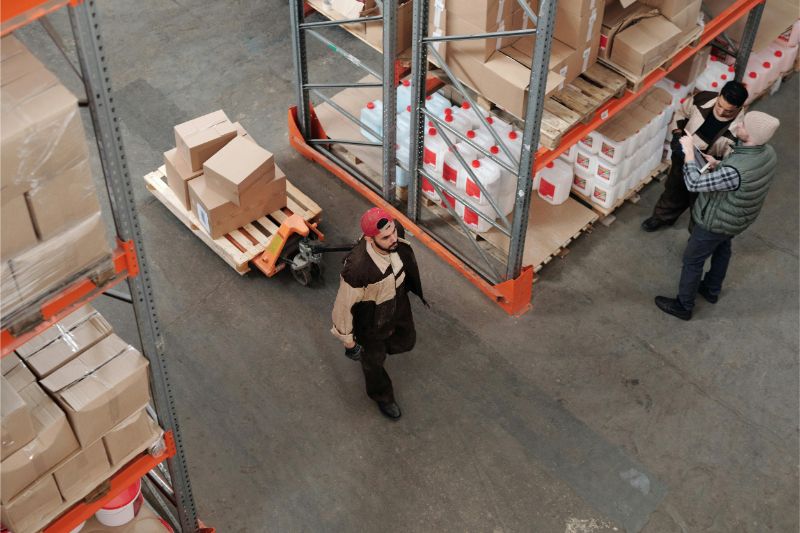 Overhead view of a busy warehouse aisle with pallet racks, stacked boxes, a worker pulling a pallet jack and two colleagues checking inventory — storage solution.