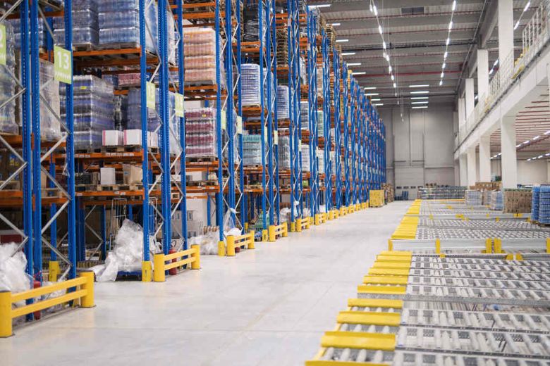 High-bay warehouse aisle with tall blue-and-orange pallet racks stocked with wrapped pallets and empty roller conveyors in the foreground — warehousing services.