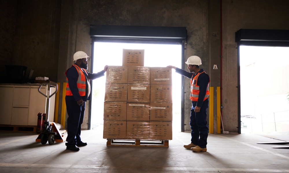Two warehouse workers wearing white hard hats and orange safety vests